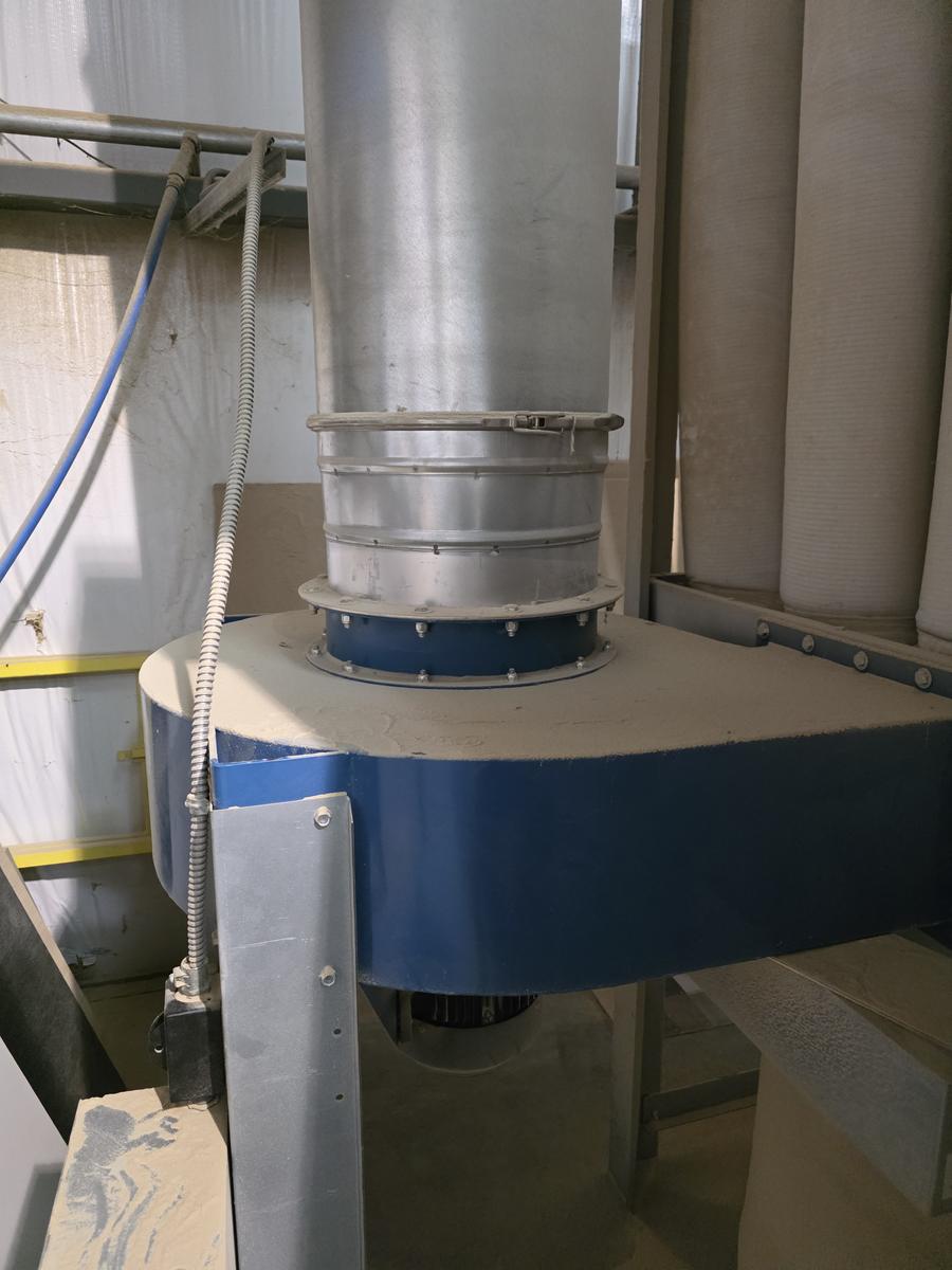 A round metal duct drops into the top of a blue fan housing on a dust collector, joined with a clamp band above a bolted flange. Fine dust covers the top of the housing, and the collector's white fabric filter bags hang to the right.