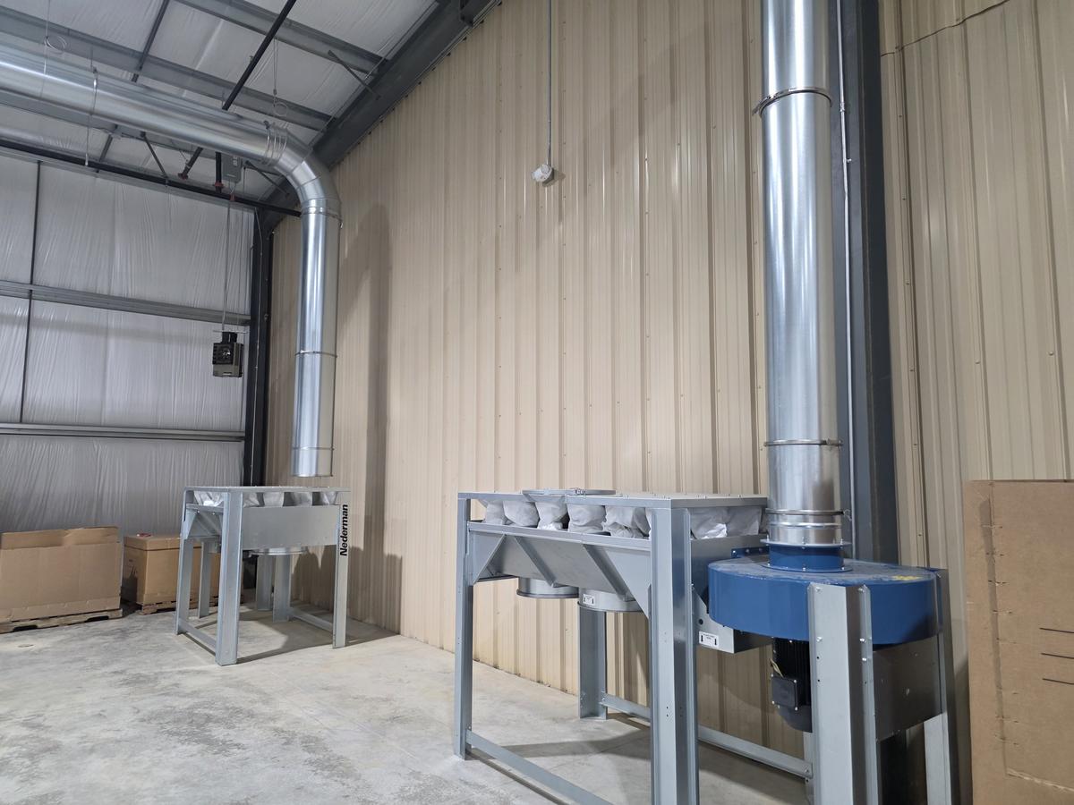 Two dust collector frames stand along a tan metal wall, one marked Nederman, with white filter bags along the top of each. A duct riser comes down from the ceiling to just above the left frame, and a second riser runs down into a blue fan housing at the end of the right frame.