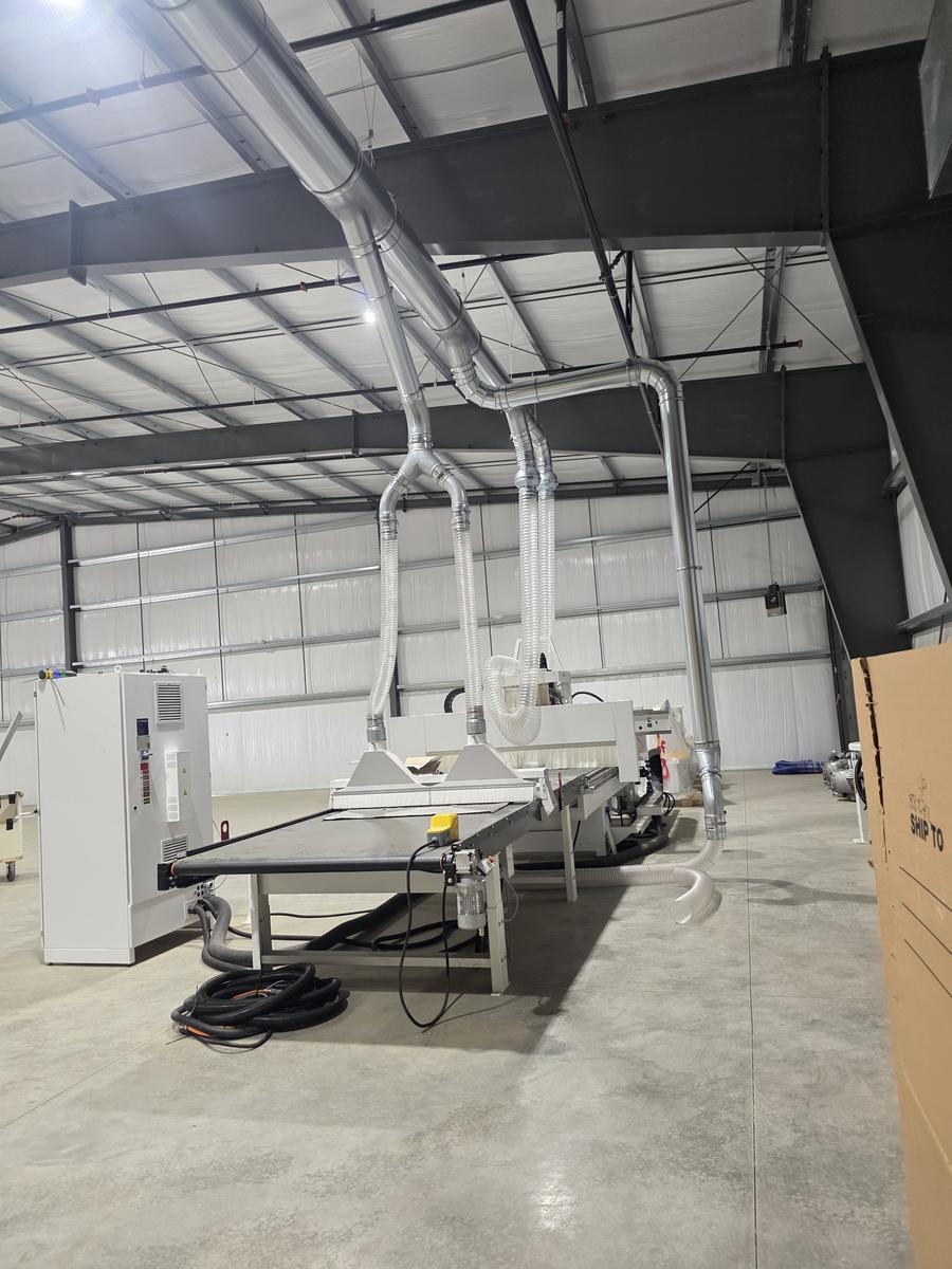 Overhead duct splits through wye fittings into drops that change to clear flexible hose and connect to the top of a large woodworking machine. Another drop runs down to floor level on the right and ends in a loop of clear hose.