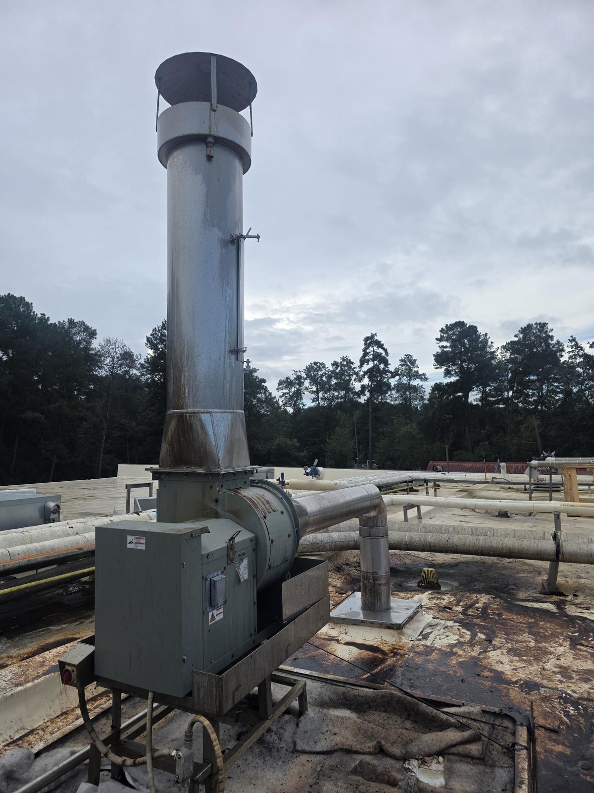 A belt-driven exhaust fan on a plant roof, connected to a tall vertical stack with a rain cap. Rust staining runs down the stack and the transition below it.