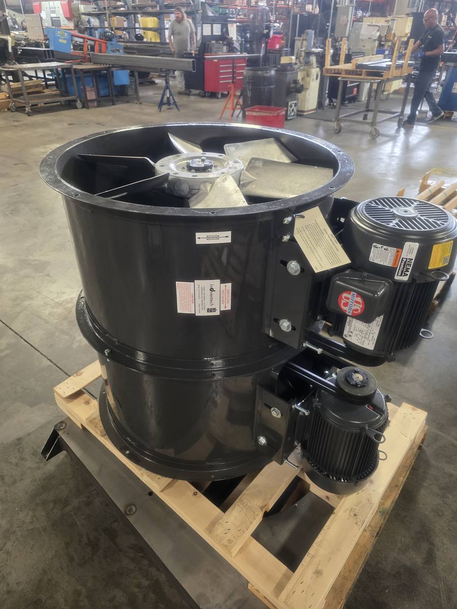 Two identical black tube-axial duct fans on a wooden pallet in a fabrication shop, each with a flanged barrel, a cast propeller and an external motor mounted on a bracket.