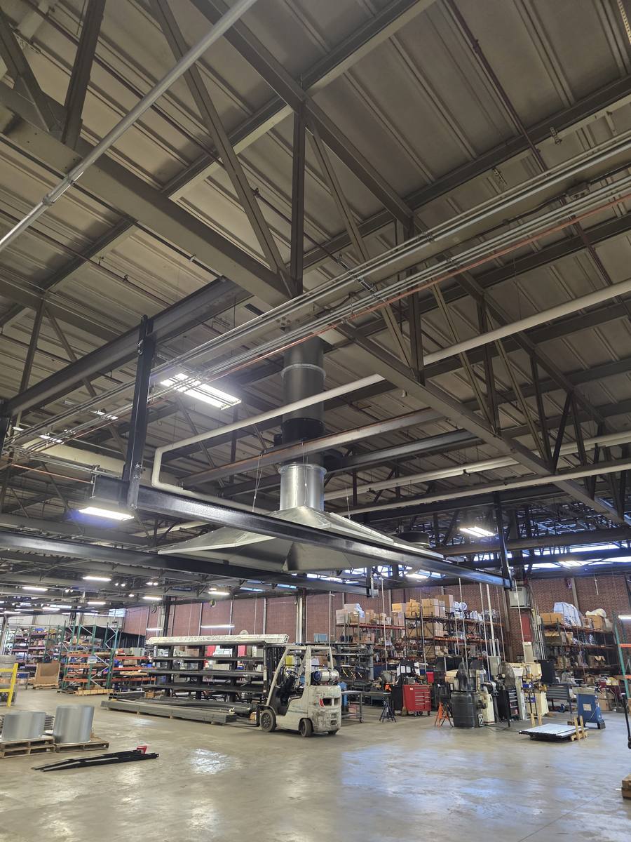 The installed capture hood: a large flat rectangular canopy suspended from the roof structure above the work area, with a round takeoff rising from its centre to the fan and the roof penetration above.
