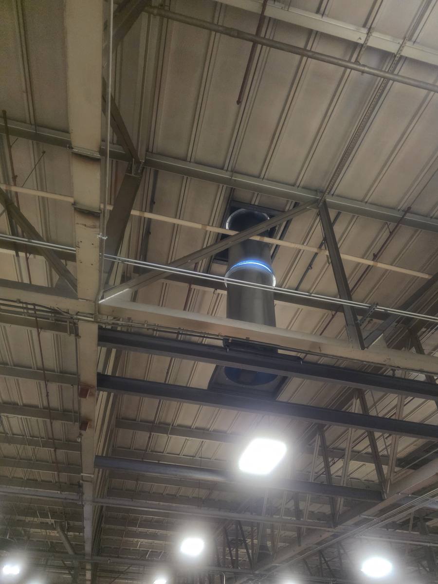 A round duct riser running vertically up through an opening in the roof deck, seen from the shop floor between steel roof joists.