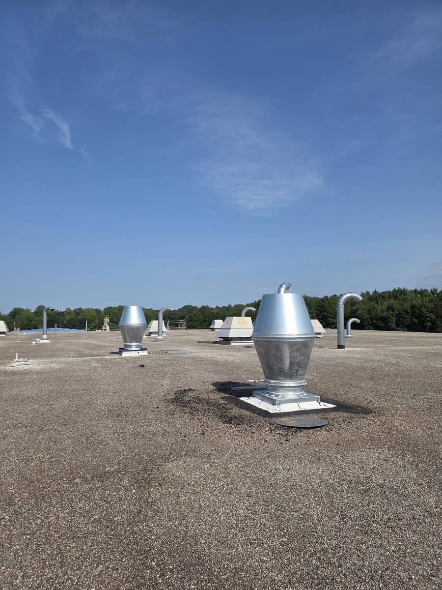 A new galvanised exhaust stack on a flat gravel roof, with a conical jet cap above a square curb cap. Older gooseneck vents and gravity ventilators stand further back under a clear sky.